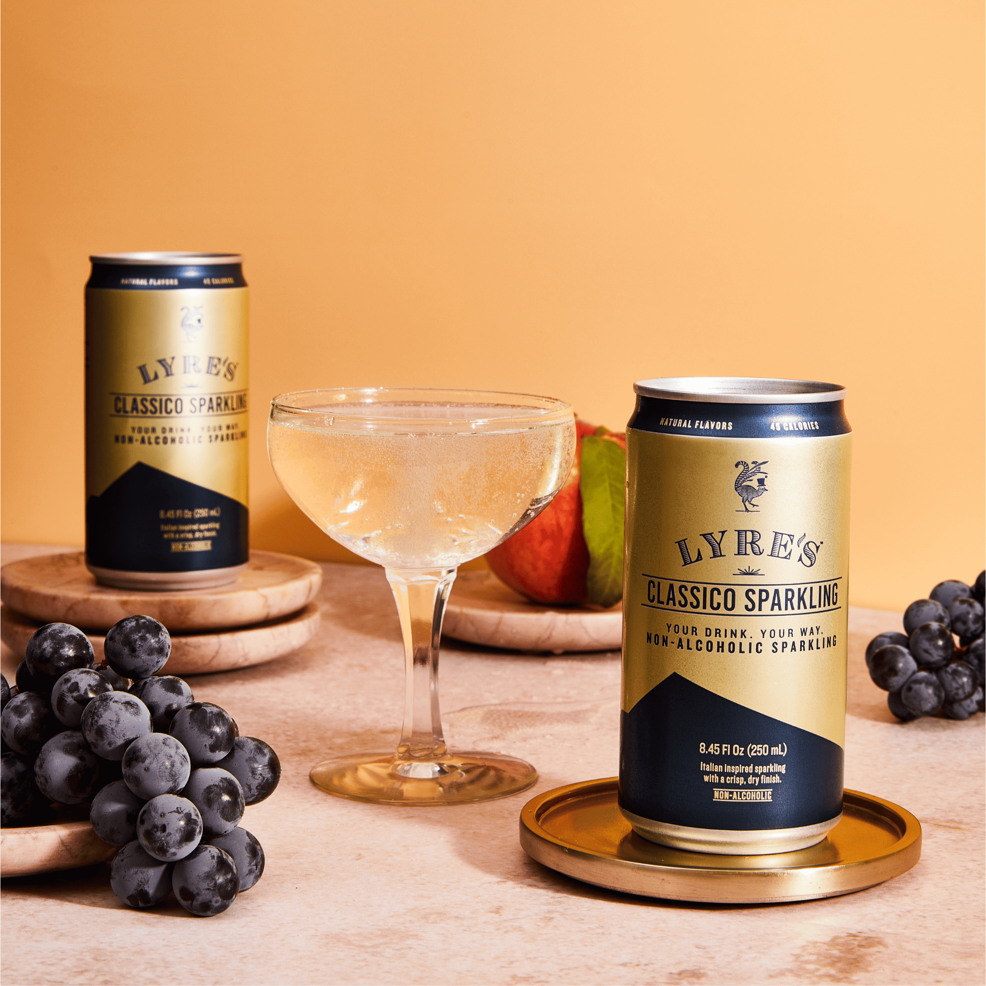Two cans of Lyre's Non Alcoholic Classic Sparkling Wine on a table with grapes and a cocktail glass.
